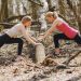 Side view of young female athletes in activewear stretching legs together using fallen tree during fitness workout in spring forest adhering to healthy lifestyle
