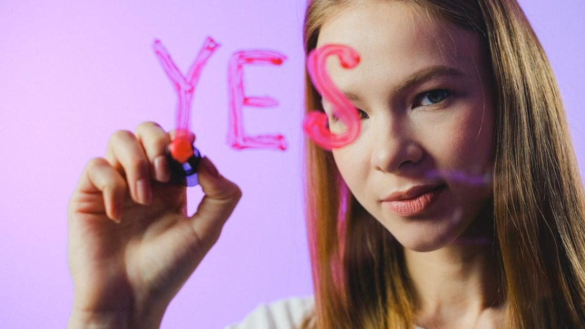 Teenage girl writing the word 'Yes' on glass with marker in studio setting.